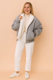 Gray bomber jacket with plush white fleece lining, styled over a beige ribbed turtleneck sweater and paired with white straight-leg pants and casual white sneakers.
