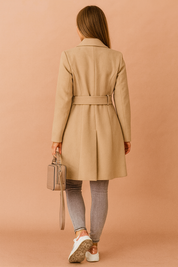 Back view of beige long coat with waist belt detail, styled over grey skinny jeans, white sneakers, and a taupe handbag.