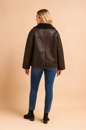 Back view of a dark brown shearling-lined jacket with wide sleeves, straight hem, and panel seam detailing, featuring a soft textured interior collar and smooth leather-look exterior.