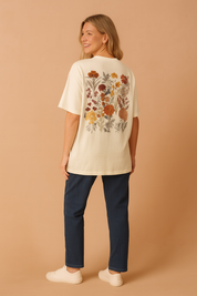 Beige short-sleeve cotton t-shirt featuring a detailed chest patch with warm-toned floral embroidery in rust, cream, and olive hues, paired with straight-cut dark brown corduroy pants and brown leather low-heeled ankle boots.