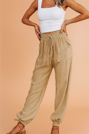 High-waisted beige drawstring linen jogger pants with front patch pockets and elasticized cuffs, styled with a fitted white asymmetrical strap crop top and tan lace-up sandals.

