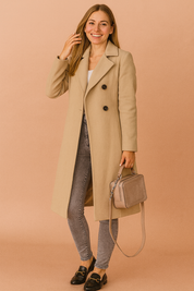 Beige long coat with notch lapels and button front, styled over a white top with grey skinny jeans, black loafers, and a taupe handbag.