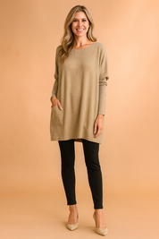 Beige oversized knit tunic with drop shoulders, long sleeves, large front patch pocket, and a boat neckline, styled with black fitted pants and neutral heels.