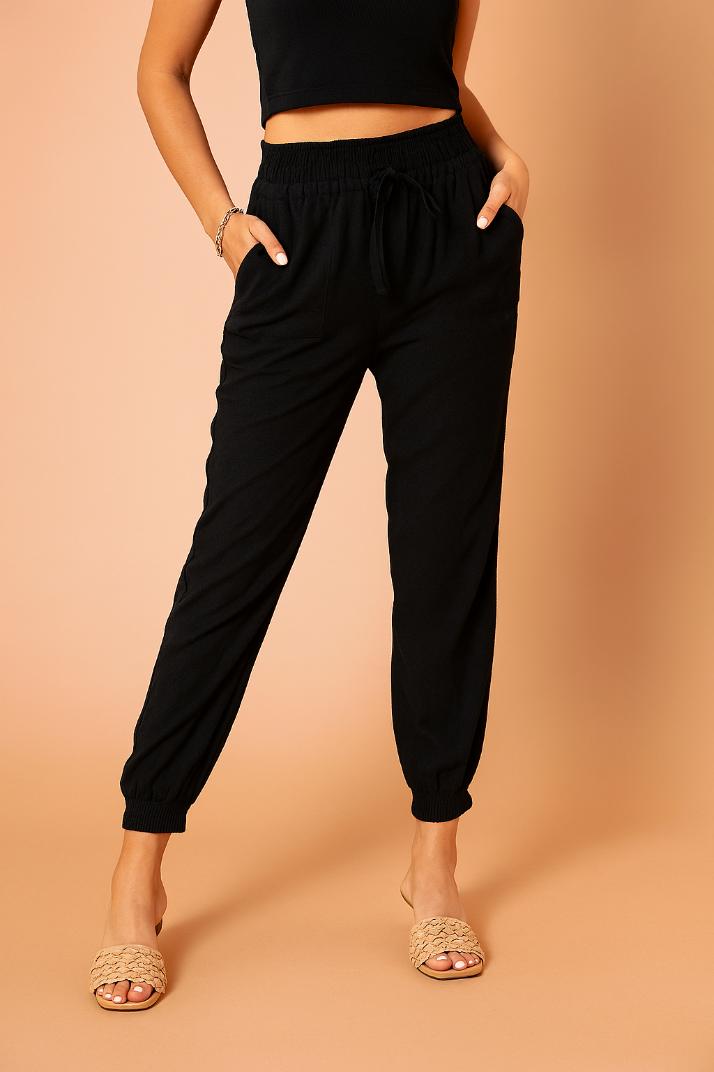 Black jogger pants featuring an elastic waistband with drawstring tie, relaxed tapered legs, side pockets, and cuffed ankles, paired with woven slip-on sandals for a laid-back yet refined look.