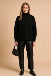 Oversized black knit turtleneck sweater with ribbed cuffs and hem, styled with straight black faux leather pants, ankle boots, and a sleek black handbag.