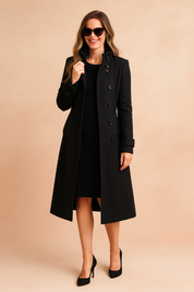 Black long trench coat with high collar, button-down front, sleeve strap detailing, and flap pockets, styled over a black dress with black high heels and sunglasses.