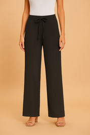 Black pleated wide-leg pants featuring a drawstring waistband and a soft ribbed texture, styled with beige heeled sandals and a tucked-in white tank top.