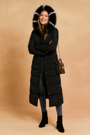 Long black quilted puffer coat with fur-trimmed hood, button-down front, paired with fitted blue jeans and black ankle boots, styled with a small crossbody handbag.