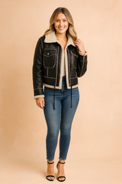 Black faux-leather shearling jacket with front pockets, beige lining, and stitched detailing, styled with slim-fit blue jeans and black heels.