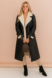 Black shearling-lined trench coat with wide lapels and double-breasted button closure, styled with beige pants, suede ankle boots, and a structured handbag.