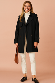 Black double-breasted trench coat with structured lapels and button front styled over a cream turtleneck and white trousers.