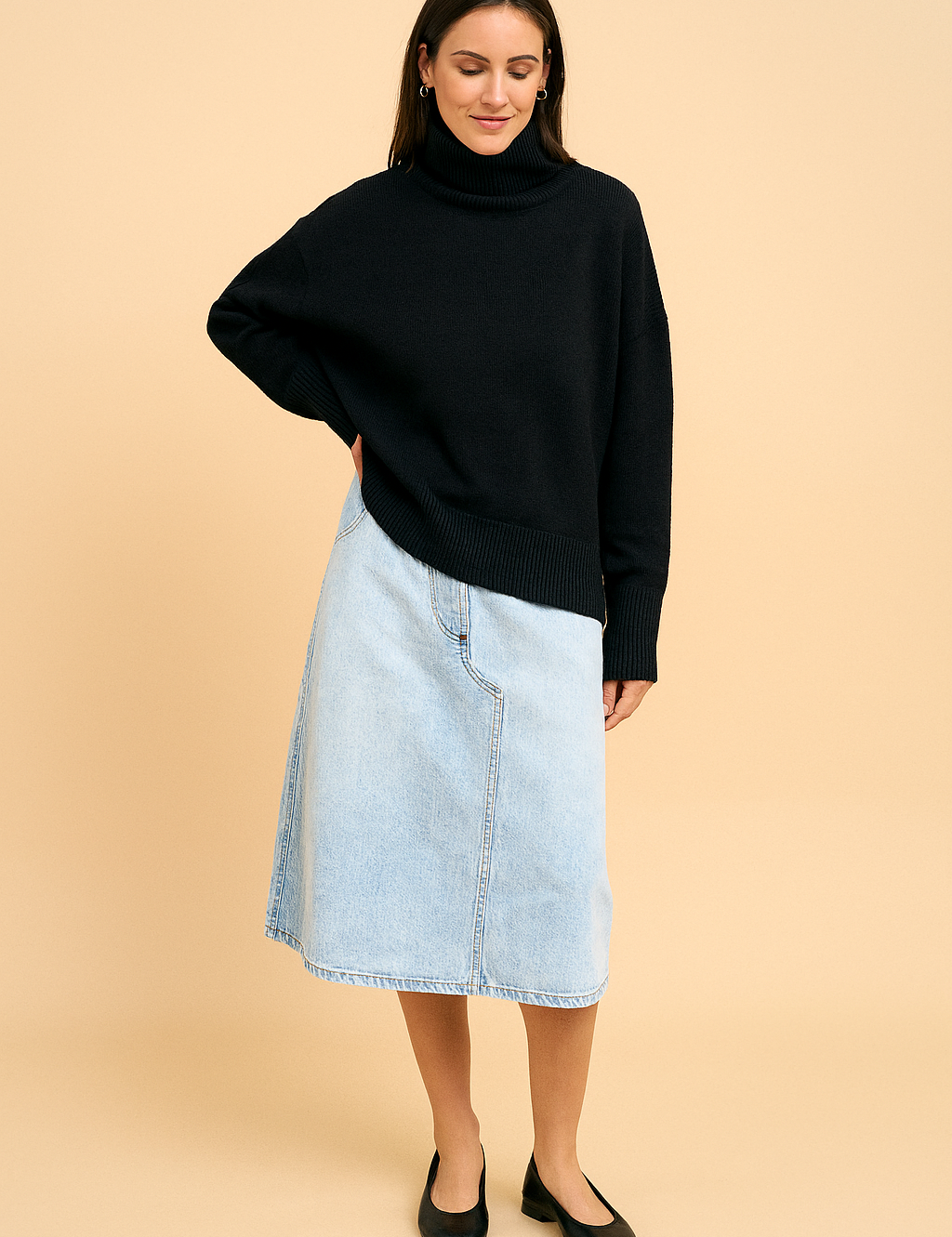 Black oversized turtleneck sweater paired with a light blue denim midi skirt and classic black ballet flats, styled with gold hoop earrings.
