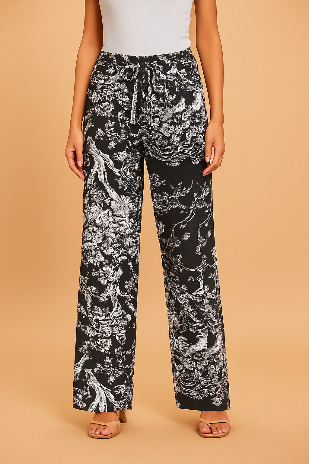 High-waisted black wide-leg pants featuring an intricate white botanical and nature-inspired print with a matching drawstring tie and button, styled with beige strappy sandals.