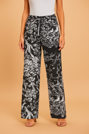 High-waisted black wide-leg pants featuring an intricate white botanical and nature-inspired print with a matching drawstring tie and button, styled with beige strappy sandals.