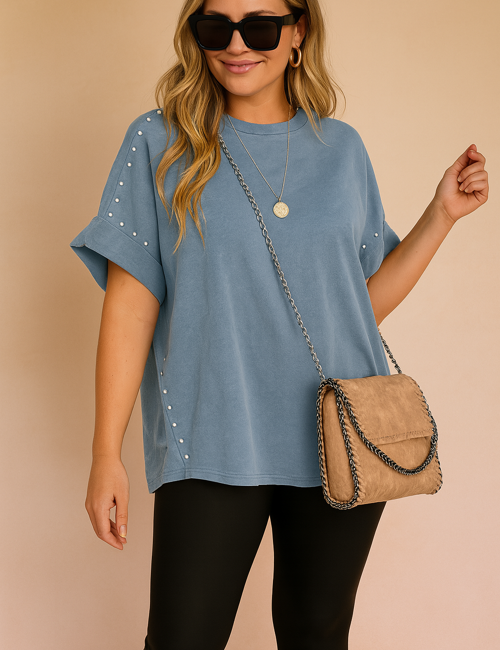 Oversized short-sleeve blue T-shirt with rolled cuffs and decorative silver studs along the sleeves and side seams, paired with black leggings and a tan suede crossbody purse with chain trim.