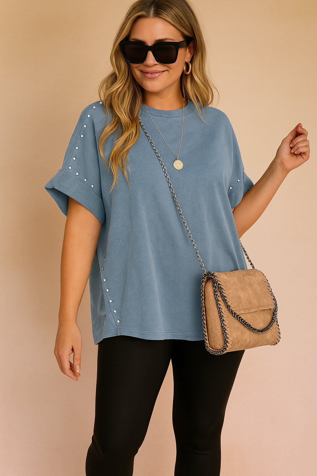 Oversized short-sleeve blue T-shirt with rolled cuffs and decorative silver studs along the sleeves and side seams, paired with black leggings and a tan suede crossbody purse with chain trim.