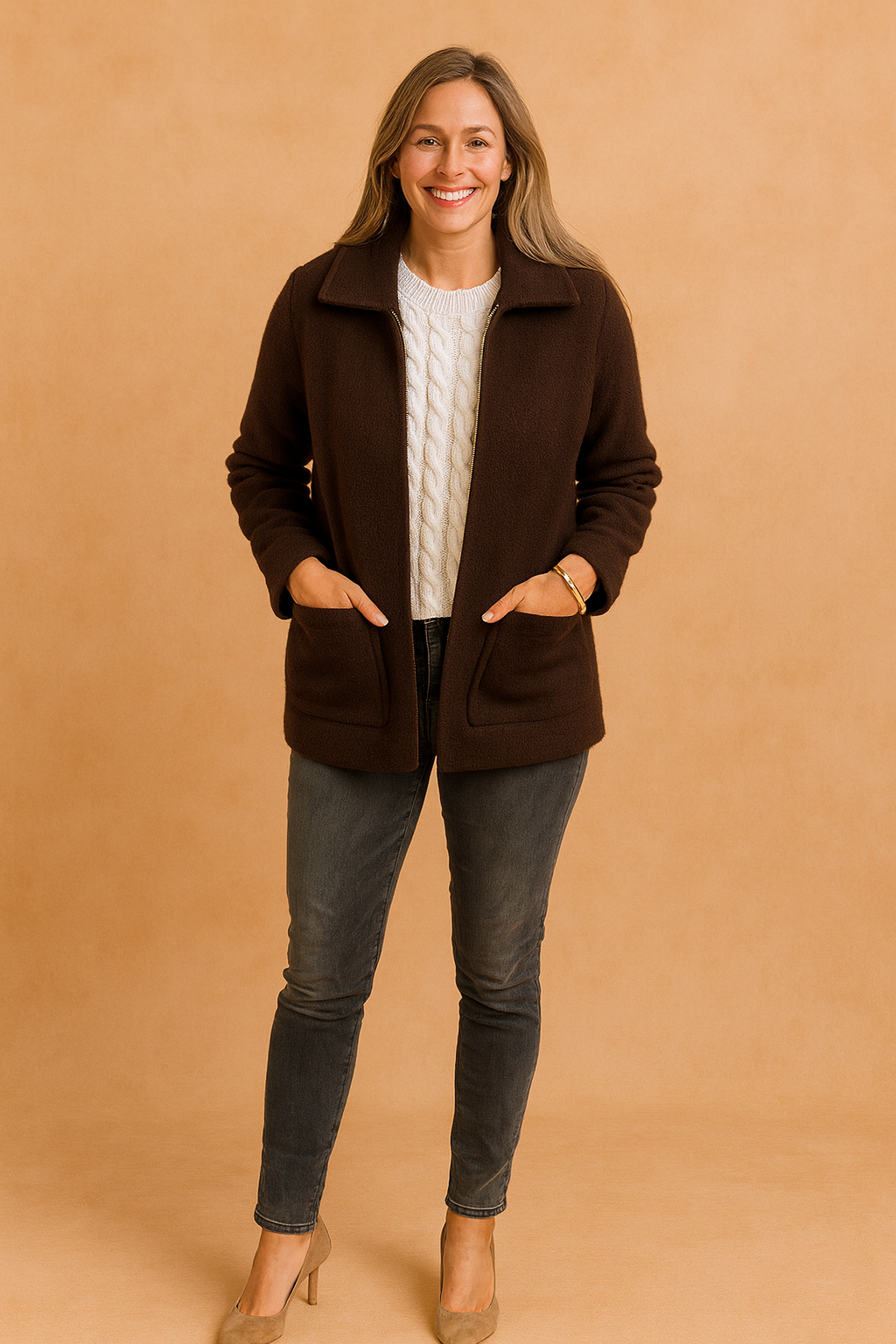 Dark brown zip-up jacket with wide collar and side pockets, layered over a white cable-knit sweater, styled with faded charcoal skinny jeans and taupe heels.