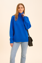 Cobalt blue oversized knit turtleneck sweater with ribbed hem and cuffs, paired with light wash straight-leg jeans and black leather shoulder bag.