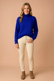Cobalt blue cable-knit high-neck sweater with ribbed cuffs and hem, paired with beige slim-fit pants and taupe ankle boots.