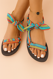 Colorful fabric strappy sandals featuring bold horizontal stripes in turquoise, red, orange, yellow, and purple, with a soft textile texture and tied ankle bows, set on black flat soles.