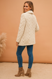 Cream textured faux shearling jacket with cable-knit pattern, featuring a soft collar and hip-length cut, paired with slim blue jeans and taupe heeled ankle boots.