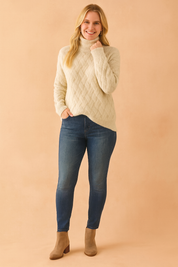 Cream cable-knit turtleneck sweater with oversized sleeves and textured diagonal pattern, paired with medium-wash skinny jeans and taupe suede ankle boots.