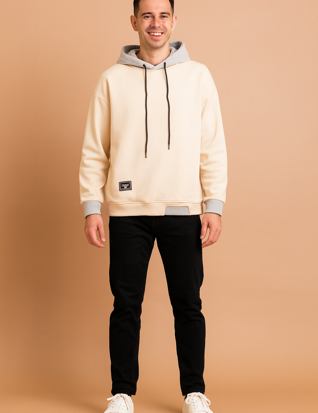 Men’s cream hoodie with contrasting gray hood lining, black drawstrings, ribbed cuffs, hem detail, and small black patch accent, styled with black slim-fit jeans and white sneakers.