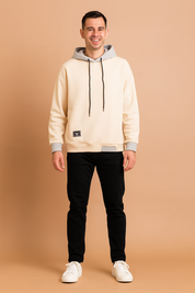 Men’s cream hoodie with contrasting gray hood lining, black drawstrings, ribbed cuffs, hem detail, and small black patch accent, styled with black slim-fit jeans and white sneakers.