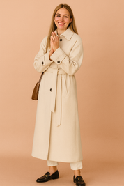 Cream long trench coat with waist tie belt, button details, and structured cuffs styled over white trousers and black loafers.