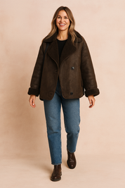 Dark brown shearling-lined aviator jacket featuring oversized lapels, double-breasted button closure, and rolled cuffs, paired with straight-cut medium blue jeans and dark leather ankle boots.