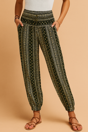 Bohemian-style harem pants in dark green with vertical geometric and floral prints, elastic ankle cuffs, and a wide smocked waistband, styled with tan strappy sandals.