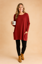 Deep red oversized tunic sweater without buttons, featuring long relaxed sleeves, front patch pockets, and soft knit texture, styled with black leggings and tan suede ankle boots.