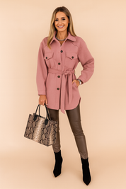 Dusty pink belted button-up jacket with flap chest pockets and black button detailing, styled with sleek leather pants, a snakeskin tote bag, and black ankle boots.