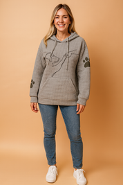 Grey waffle-knit hoodie featuring a minimalist dog and hand line art on the chest, paired with black paw print graphics on the sleeves; the top includes a front kangaroo pocket and drawstring hood, styled with light blue slim-fit jeans and white sneakers.