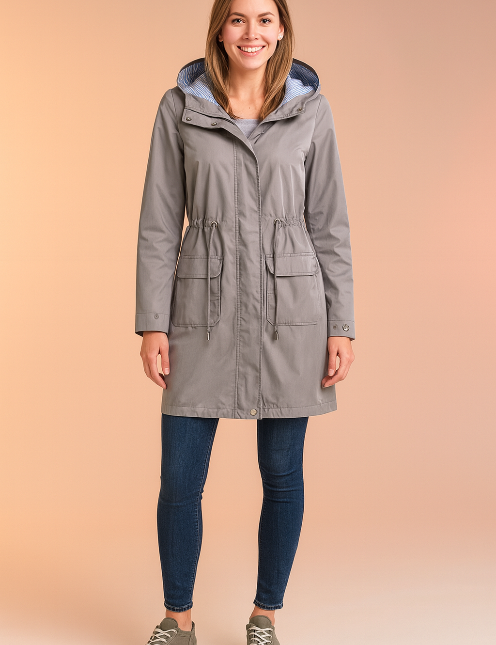 Light grey hooded raincoat with striped inner lining, front drawstring waist, snap-button closure, and two large flap pockets, styled with casual sneakers and fitted jeans.