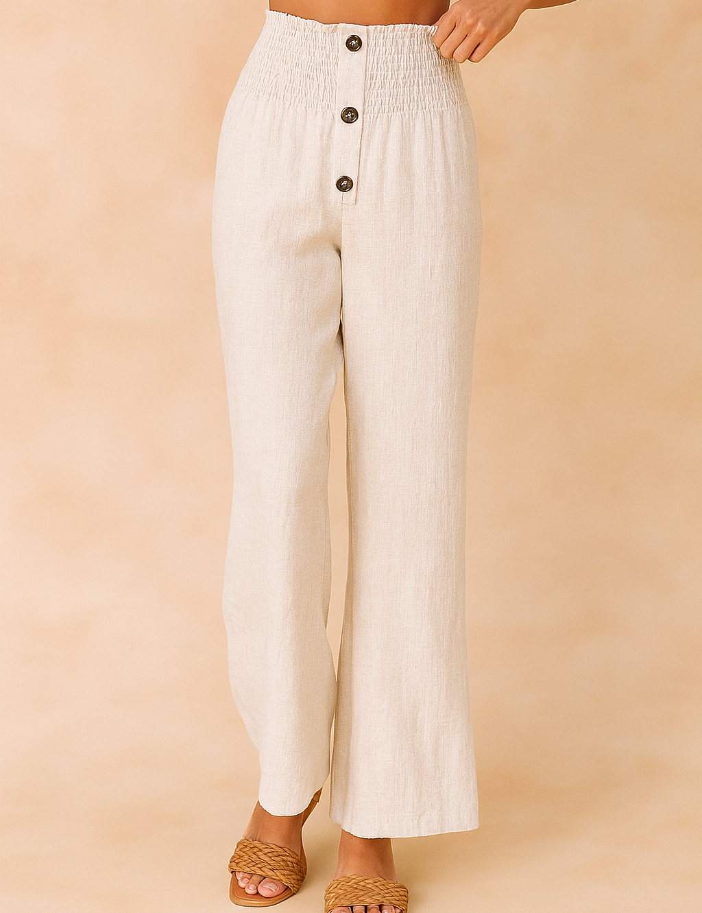 High-waisted off-white linen pants with a smocked waistband, four brown buttons, and a wide-leg cut, styled with woven tan sandals and a white crop top.
