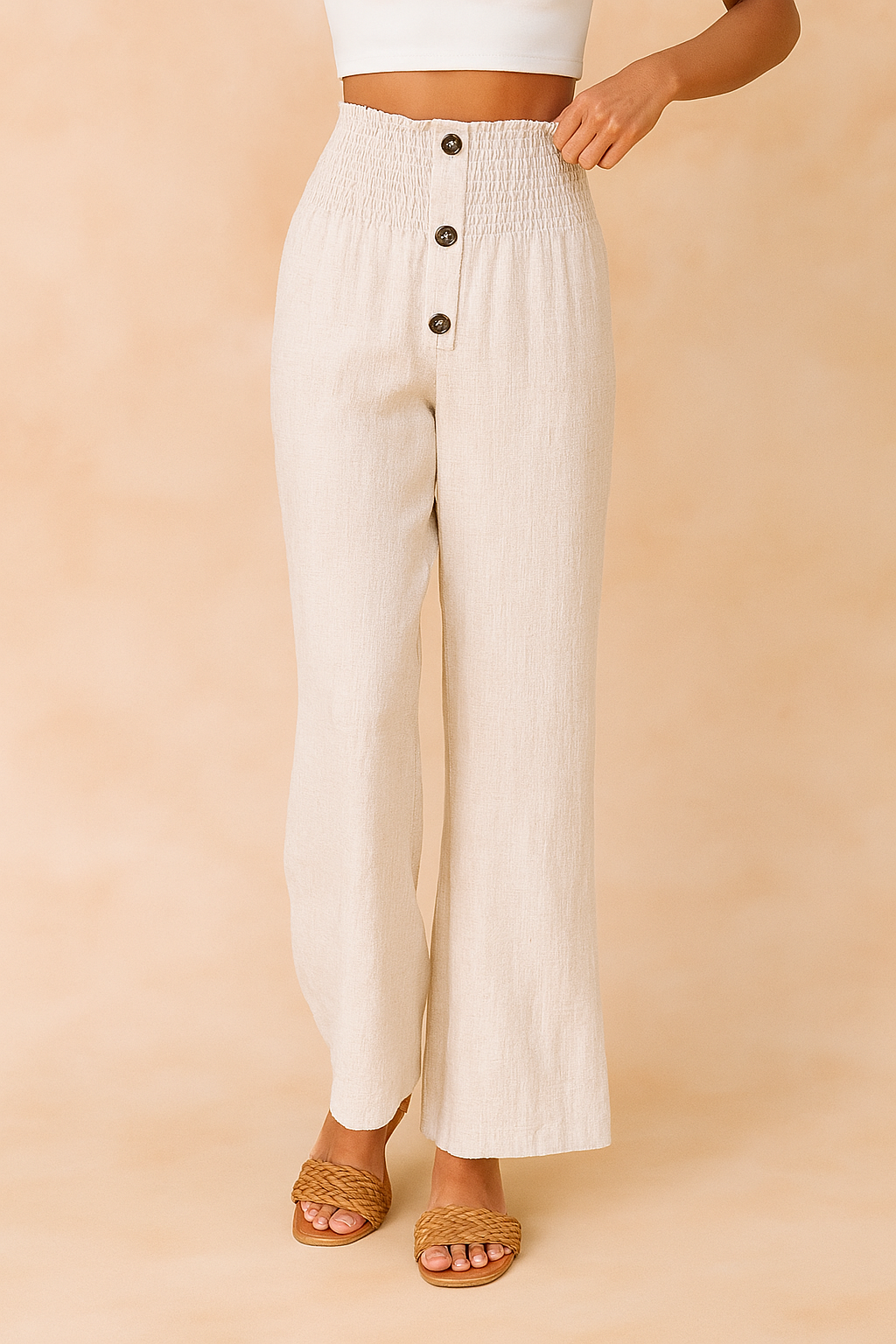 High-waisted off-white linen pants with a smocked waistband, four brown buttons, and a wide-leg cut, styled with woven tan sandals and a white crop top.