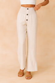 High-waisted off-white linen pants with a smocked waistband, four brown buttons, and a wide-leg cut, styled with woven tan sandals and a white crop top.