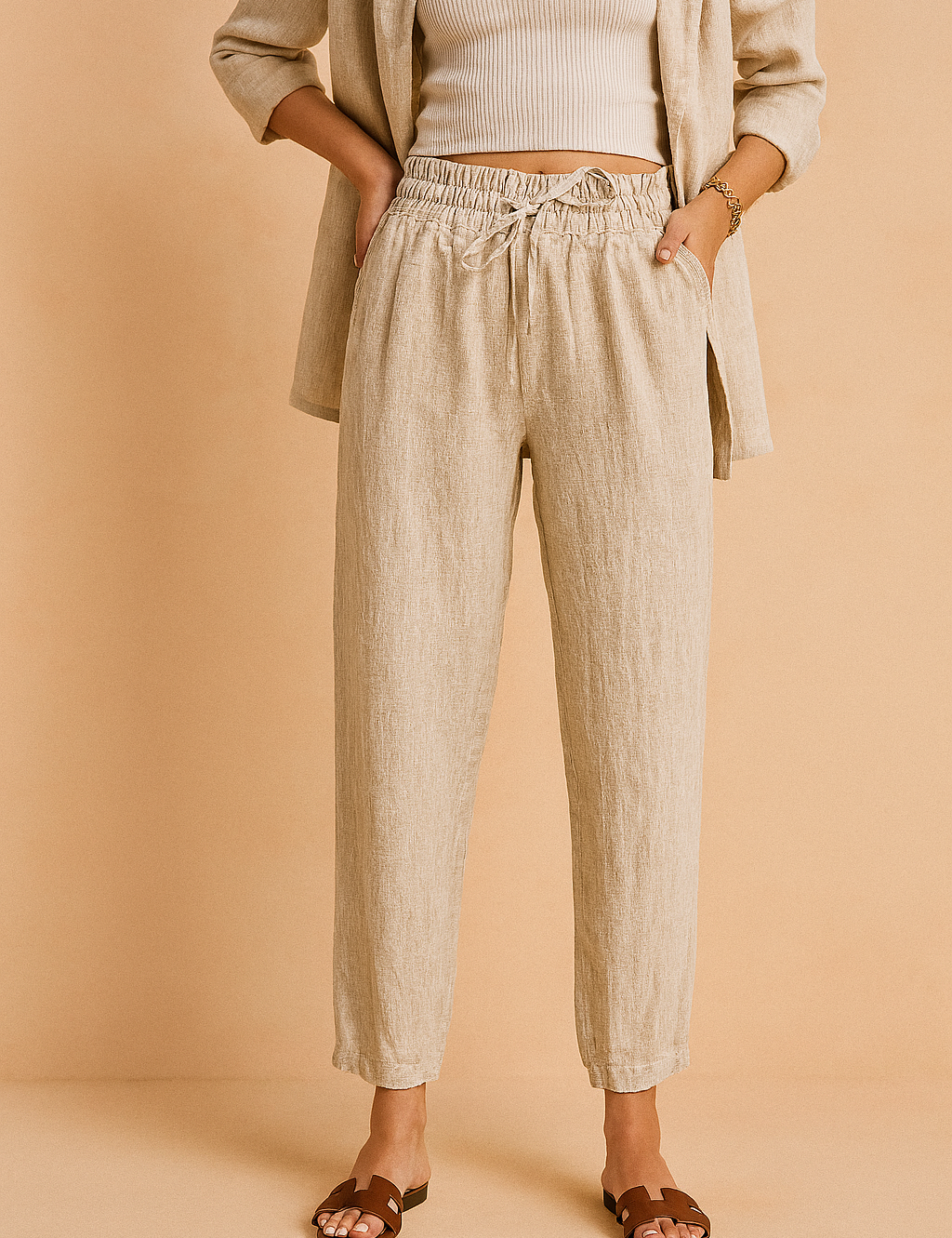 High-waisted beige linen drawstring pants with a tapered ankle-length cut, styled with a matching relaxed blazer and brown slide sandals for a polished casual look.