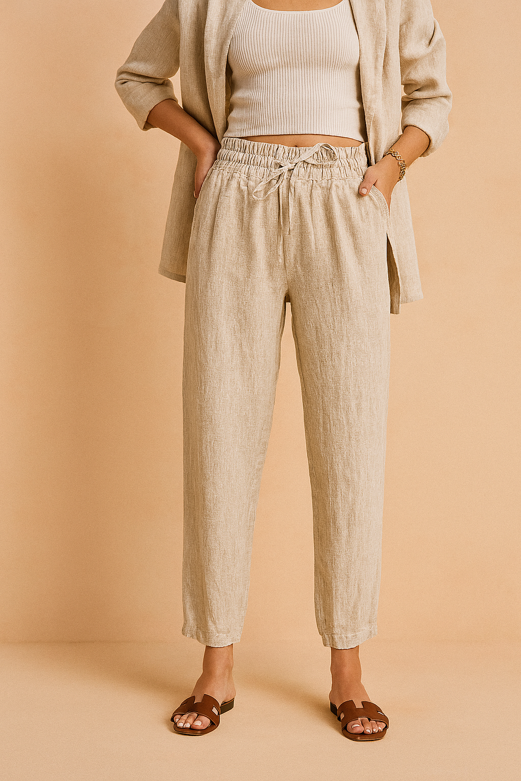 High-waisted beige linen drawstring pants with a tapered ankle-length cut, styled with a matching relaxed blazer and brown slide sandals for a polished casual look.