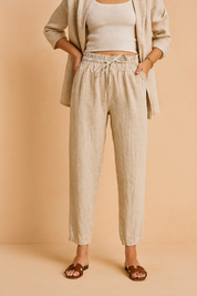 High-waisted beige linen drawstring pants with a tapered ankle-length cut, styled with a matching relaxed blazer and brown slide sandals for a polished casual look.