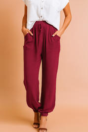 High-waisted burgundy drawstring jogger pants with front patch pockets and elastic ankle cuffs, styled with a short-sleeve white button-up blouse and brown open-toe heels.