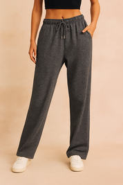 Dark charcoal fleece lounge sweatpants with a straight-leg cut, elastic waistband, matching drawstring with silver aglets, and side pockets, styled with casual off-white sneakers.