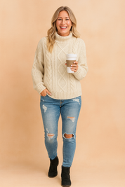 Ivory cable knit sweater with high neckline, intricate braided and diamond knit patterns, loose fit with ribbed cuffs and hem, styled with distressed denim jeans.