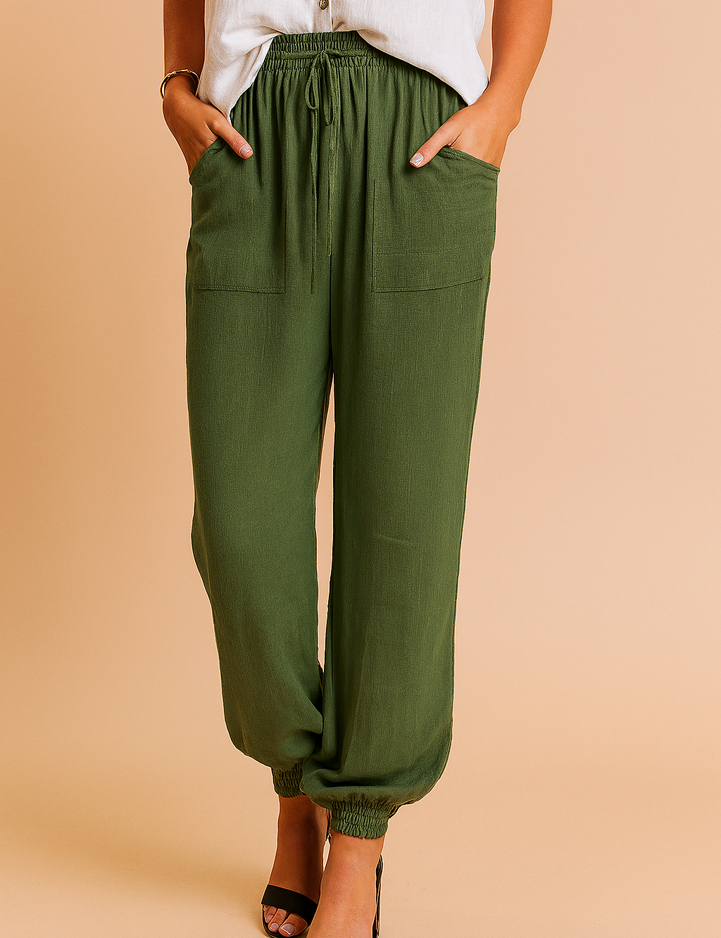 Olive green jogger-style linen pants with a drawstring waistband, oversized front patch pockets, and elasticated cuffs, styled with open-toe heels and a sleeveless off-white blouse.