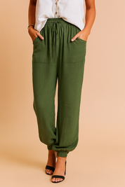 Olive green jogger-style linen pants with a drawstring waistband, oversized front patch pockets, and elasticated cuffs, styled with open-toe heels and a sleeveless off-white blouse.