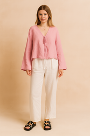 Pastel pink tie-front blouse with long sleeves styled with relaxed white pants and black daisy-embellished sandals, perfect for a light summer outfit.