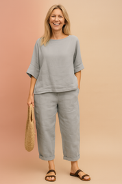 Two-piece light grey linen set with loose half-sleeve top and relaxed ankle-length pants, styled with a rolled hem and matched with brown flat sandals and a woven straw bag.