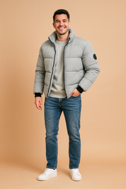 Light grey padded puffer jacket with high collar, black zipper, ribbed cuffs, and circular patch on the arm, paired with casual jeans and white sneakers.