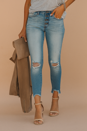 High-waisted light-wash distressed skinny jeans with exposed button-fly closure, frayed ankle hems, and knee rips, styled with taupe ankle-strap heels and a neutral-toned outerwear piece.